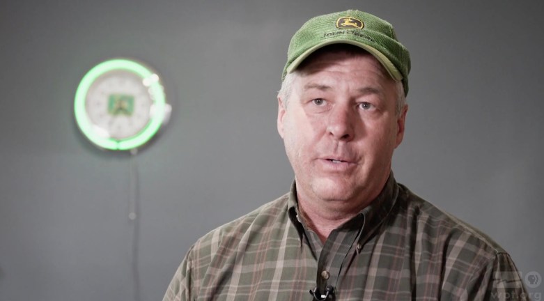 Jeff Sauer in a plaid shirt and green John Deere hat with a green-bordered circular clock in the background