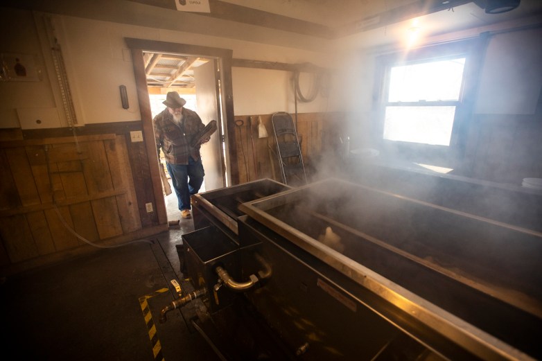 A man in a brown hat and brown coat carries firewood into a room where steam is wafting as maple syrup boils in a tank.