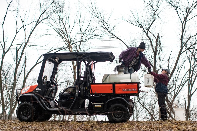 A worker hands a bucket to another worker on a motorized cart with a tank in the back.
