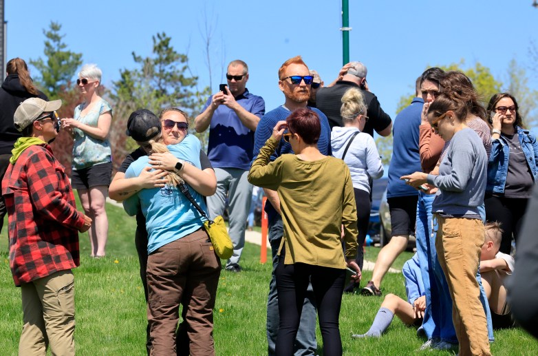 Two people hug as people gather on grass under a blue sky.