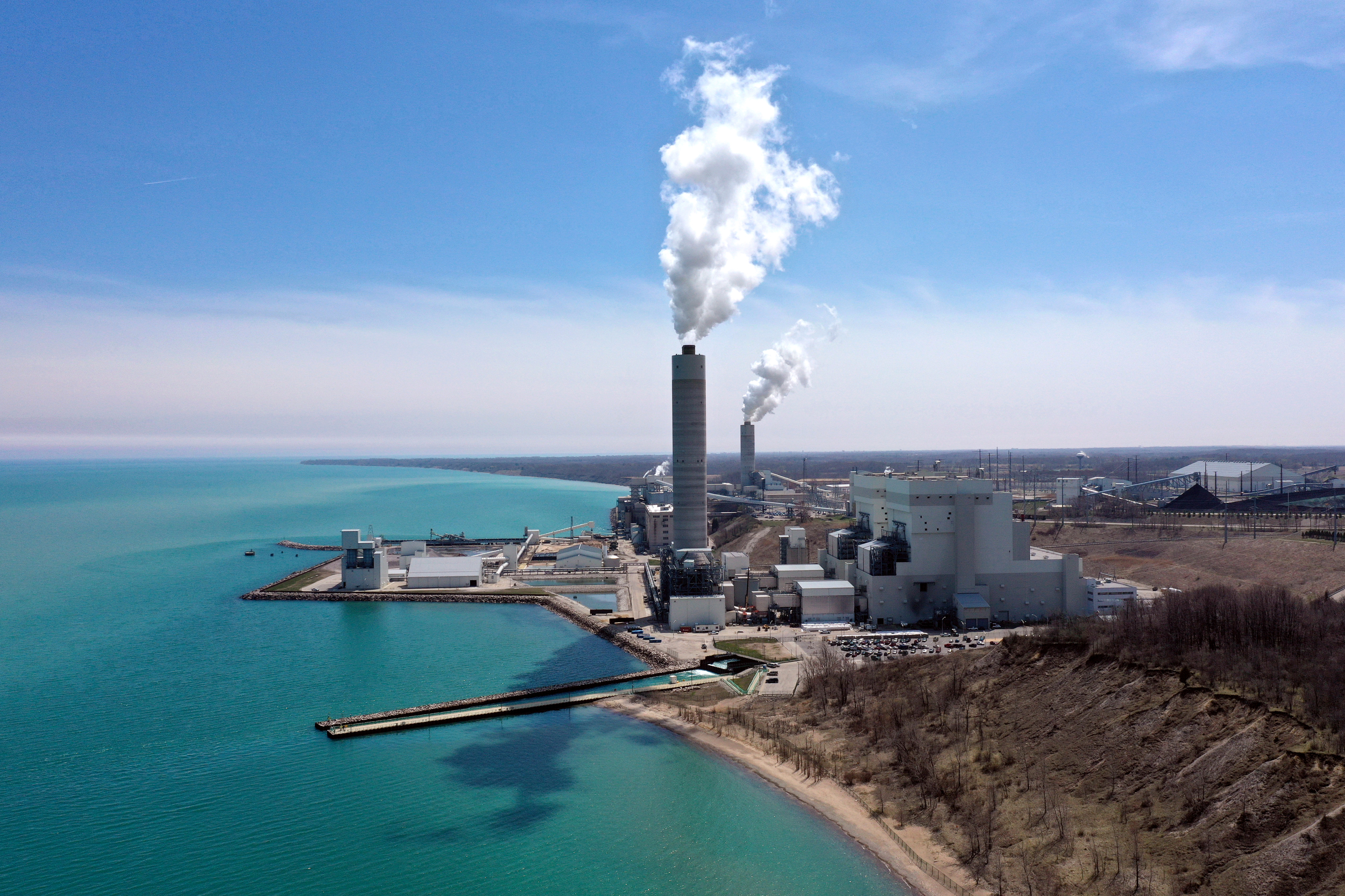 Two smoke plumes billow into a blue sky at a power plant next to a lake.