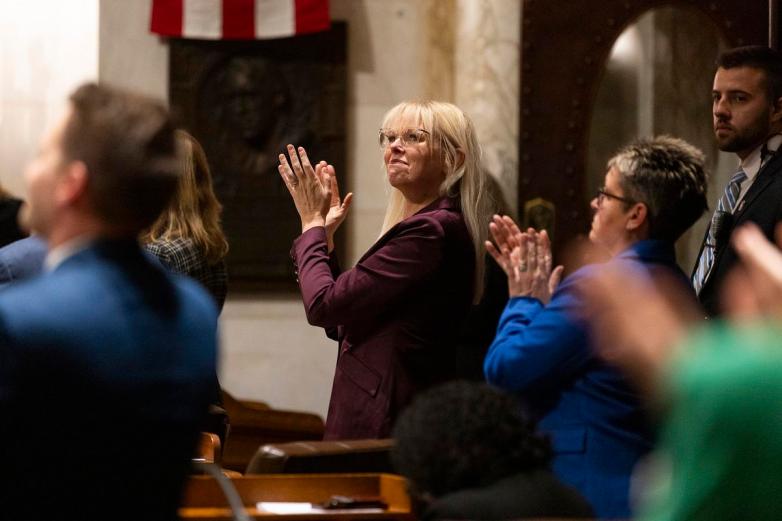 People stand and clap in a room, with a person in a blazer clapping in the center.