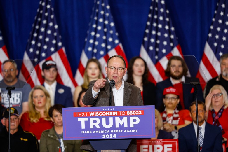 A person speaks at a podium with a sign reading "TRUMP MAKE AMERICA GREAT AGAIN! 2024" and "TEXT WISCONSIN TO 88022," with U.S. flags and people in the background.