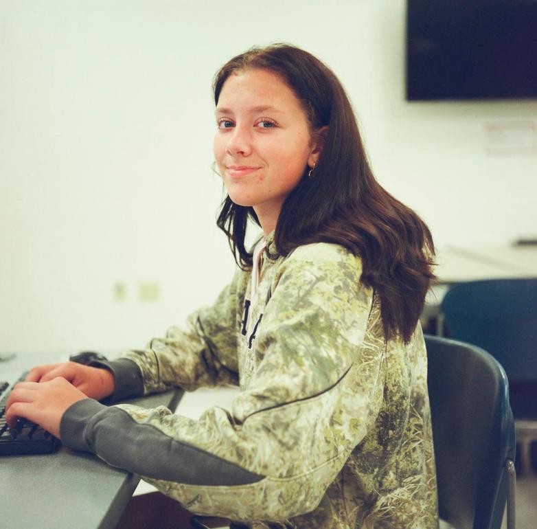 A person wearing a camouflage-patterned sweatshirt sits on a chair next to a table with hands over a computer keyboard and looks toward the camera.