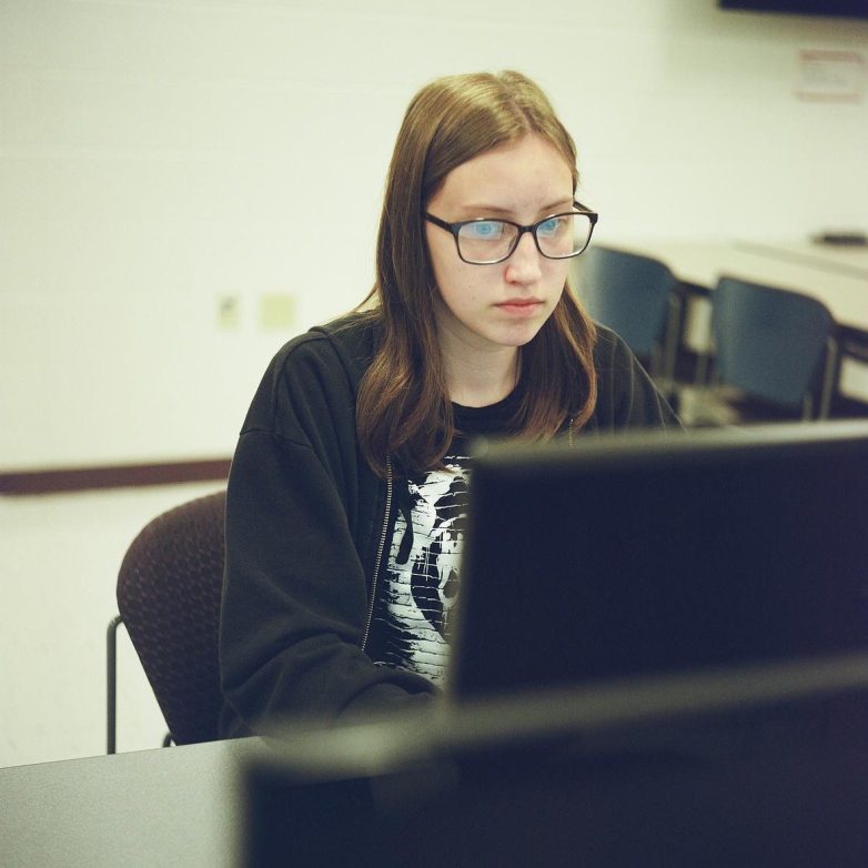 A person wearing glasses and a dark jacket looks at a computer in a room with empty chairs and tables in the background.