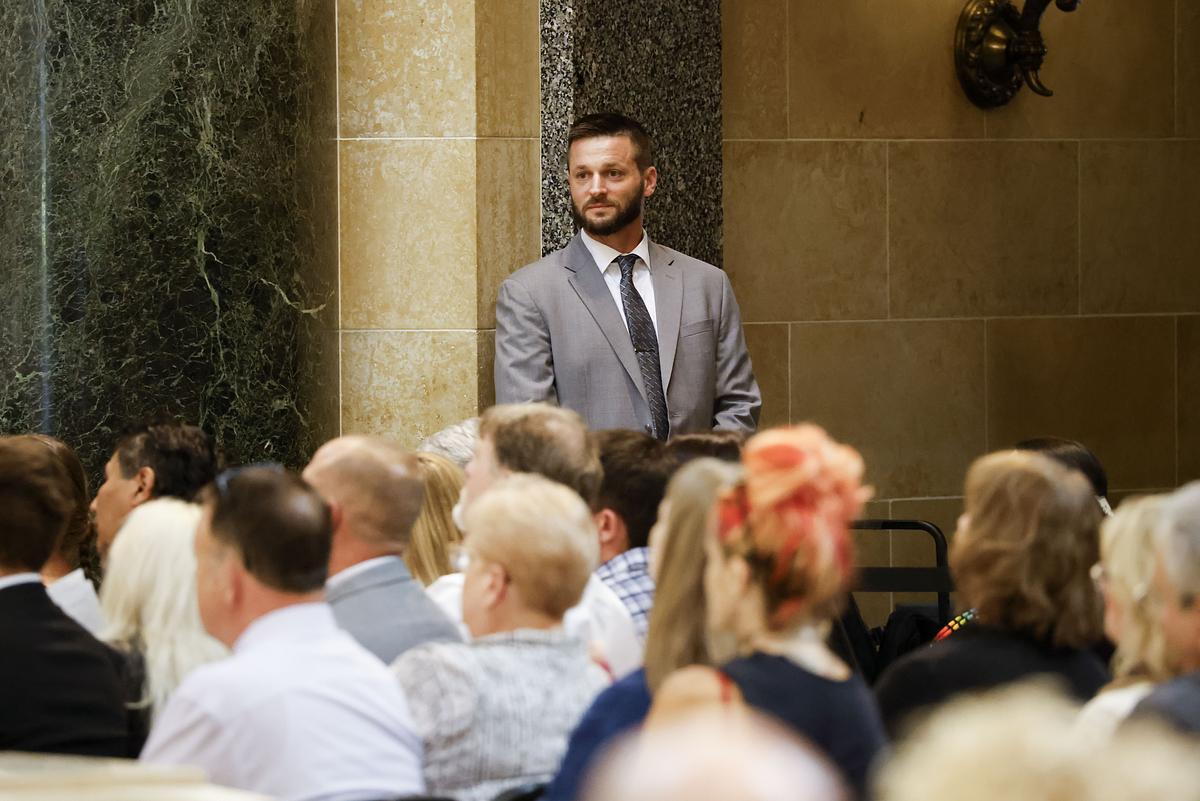 A person in a gray suit stands against a marble and stone wall while people who are sitting face the other way in the foreground.