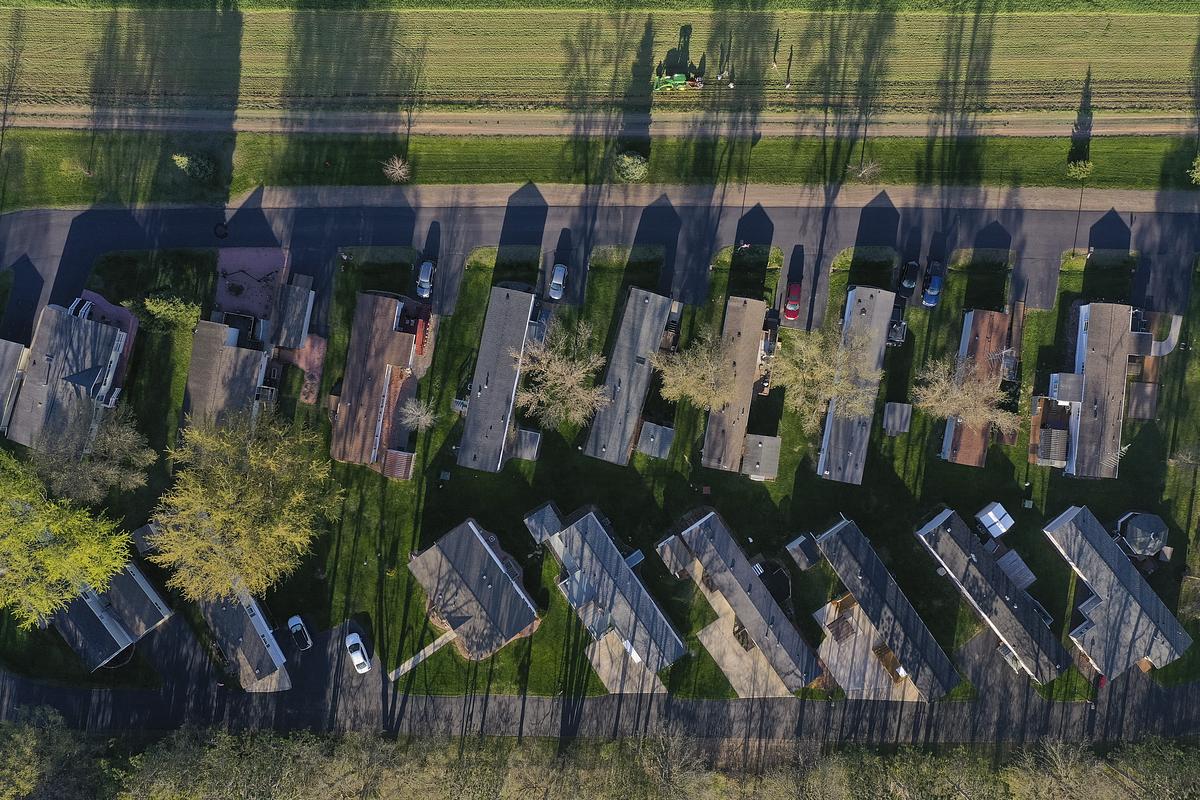 Aerial view shows rows of homes with driveways, parked vehicles and green lawns bordered by roads and fields.