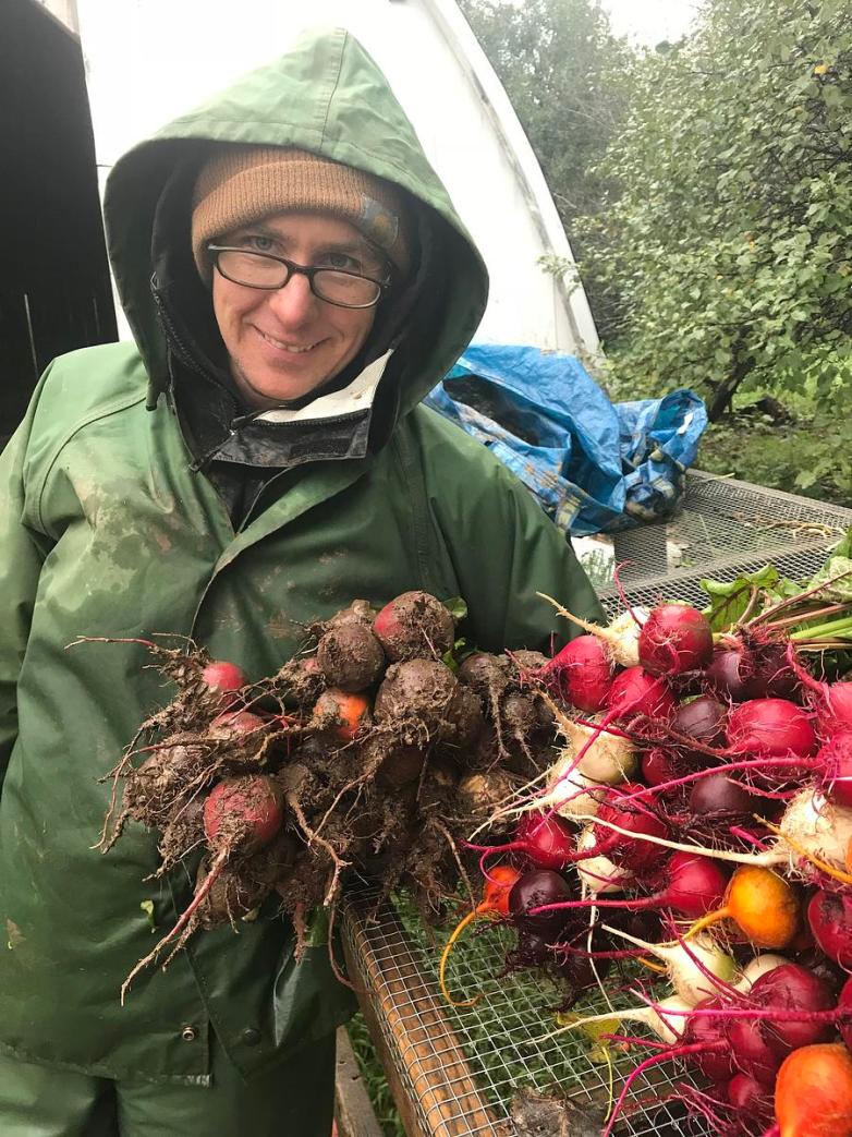 A person wearing a green rain jacket, tan knit hat and glasses holds a bunch of beets covered in soil beside a table with more cleaned vegetables.