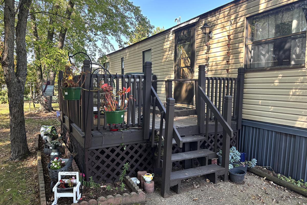 A tan mobile home with a dark brown wooden porch with potted plants and hanging baskets surrounded by trees and decorations.