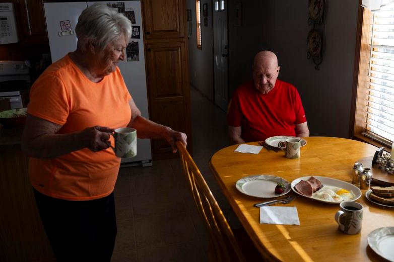 A person in an orange shirt holds a mug while standing near a wooden table set with plates of food. Another person sits nearby.