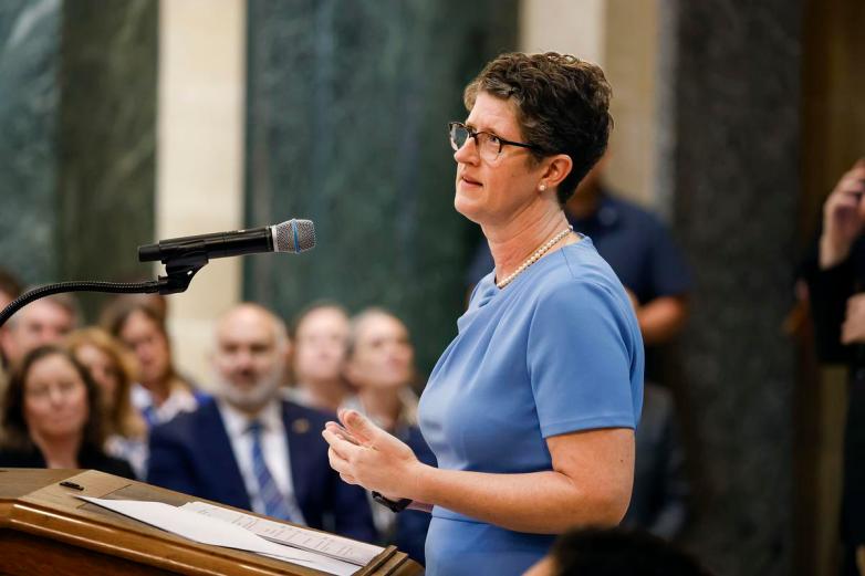 A person wearing glasses and blue clothing is at a podium with a microphone with other people in the background.