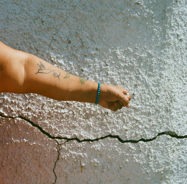 An arm with tattoos, a blue beaded bracelet and a closed fist is in front of a cracked white textured wall.