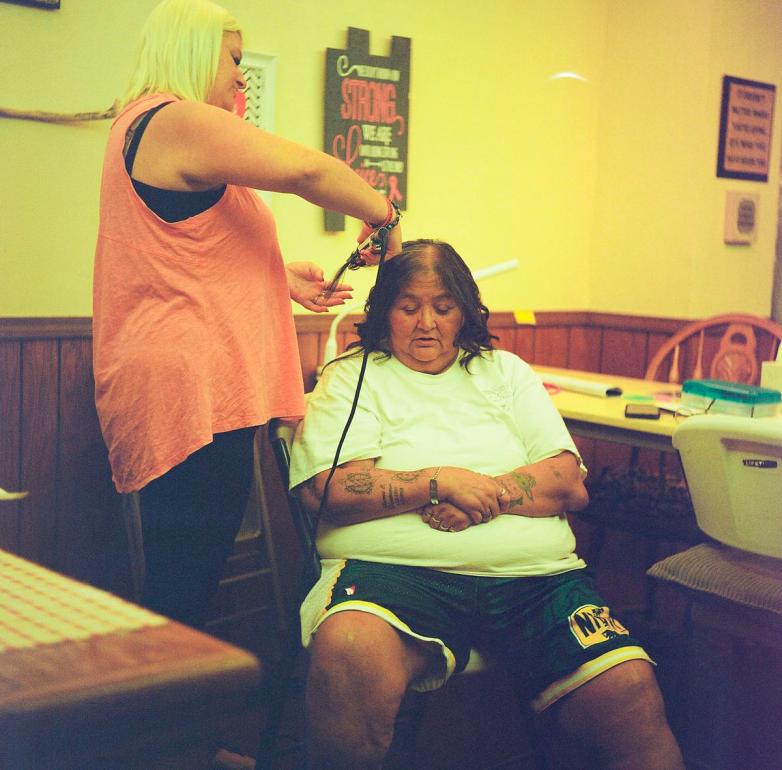 A person who is standing holds a curling iron and curls the hair of a person who is sitting in a chair in a room with wooden paneling and a yellow wall.