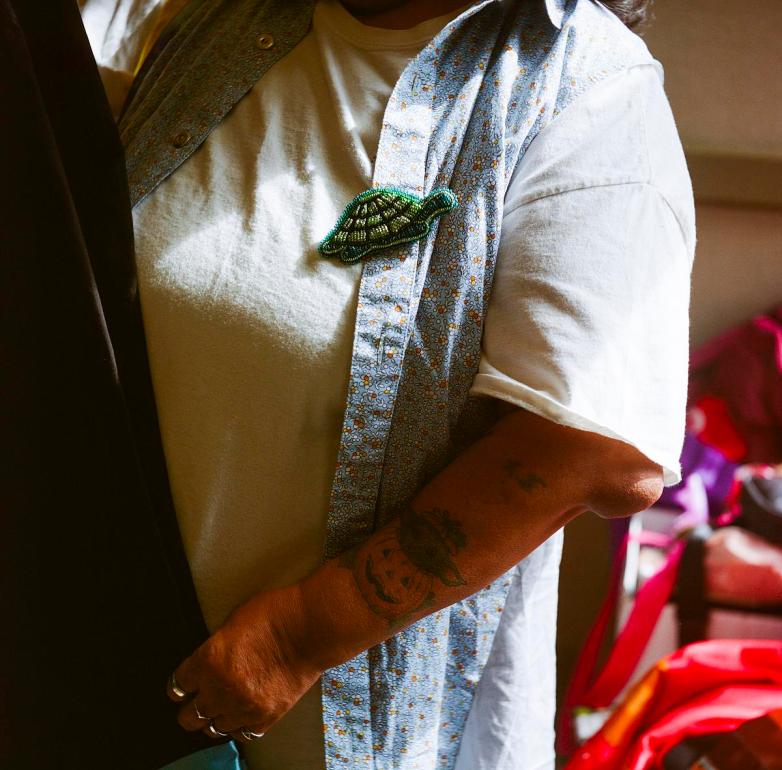 Close-up of a person wearing a white shirt and patterned vest with a green beaded turtle decoration and tattoos on the person's arm