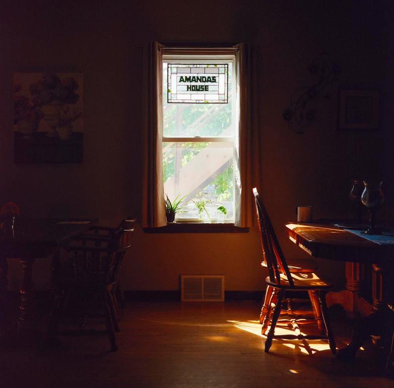Sunlight shines onto wooden chairs and a table through a window with a stained glass panel reading 