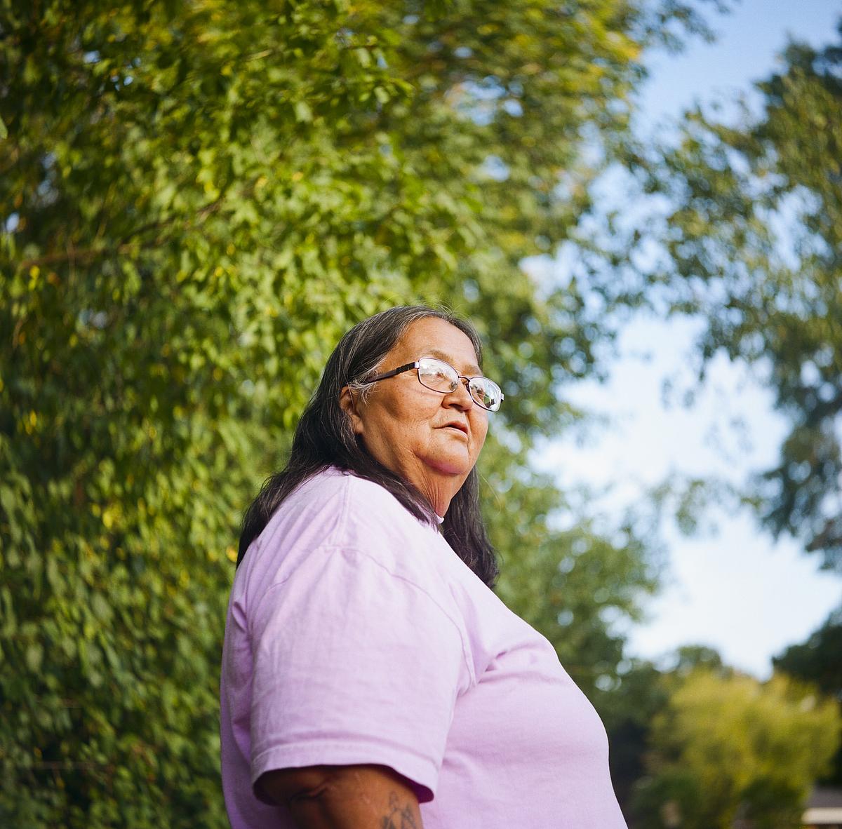 A person wearing glasses and a light purple shirt stands outdoors with trees and blue sky in the background.