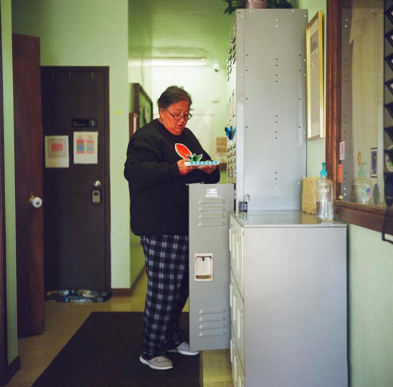 A person stands in a hallway wearing plaid pants and a dark sweatshirt while holding a pill organizer in front of an open locker.