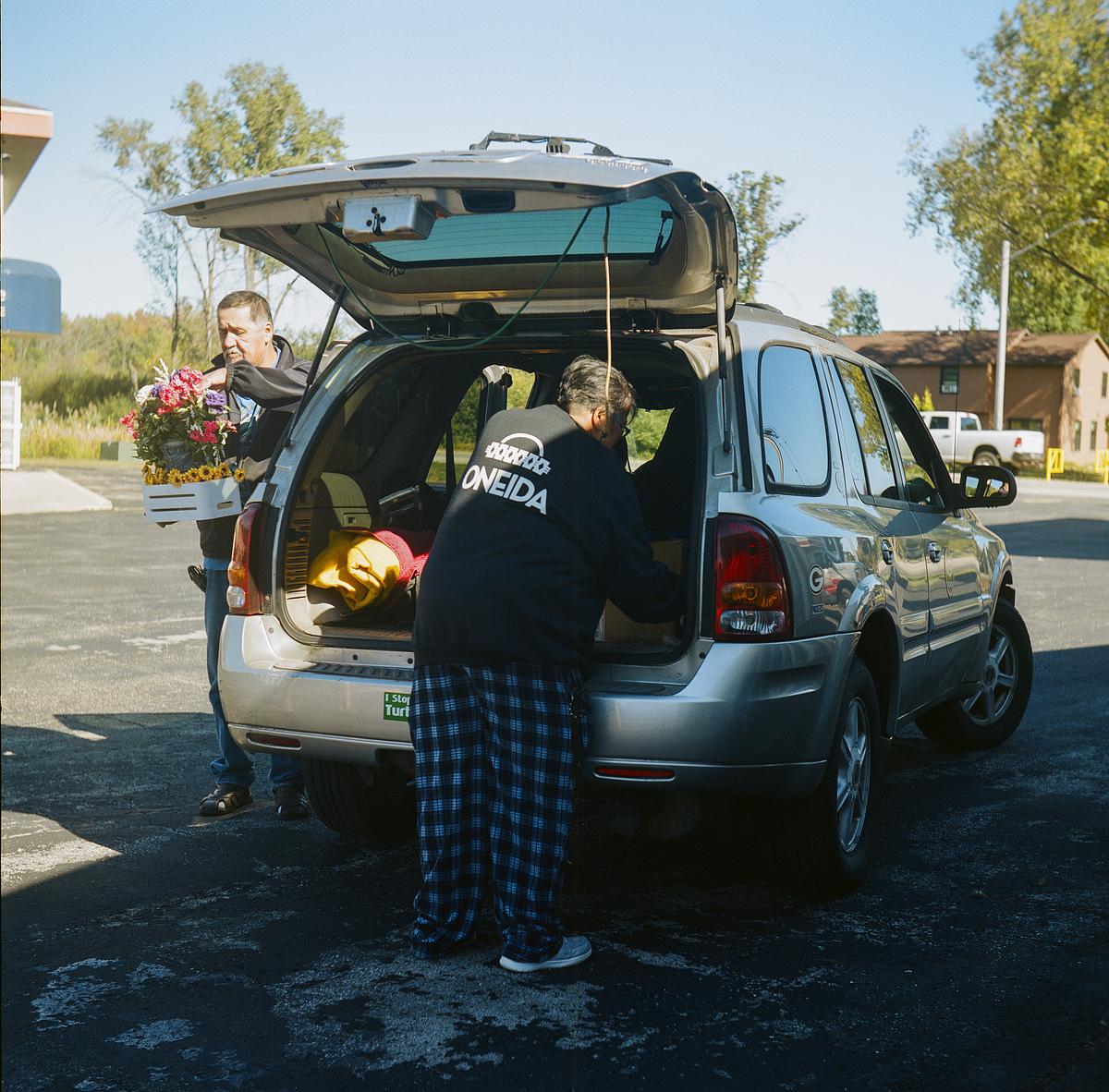 Two people load items into the back of an SUV, one holding a crate of flowers and the other wearing a top with 