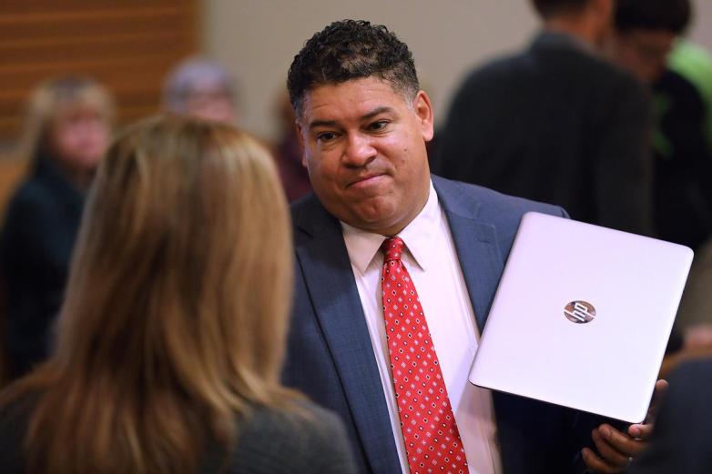 A person wearing a blue suit coat and red tie holds a silver laptop while looking at another person, with other people out of focus in the background.