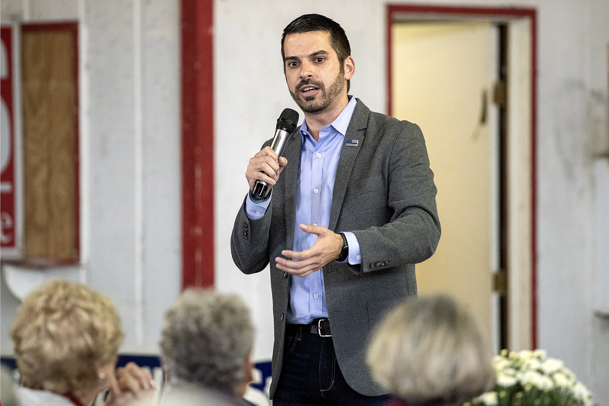 A person wearing a blazer and holding a microphone stands facing people who are seated in a room with white walls with red trim.