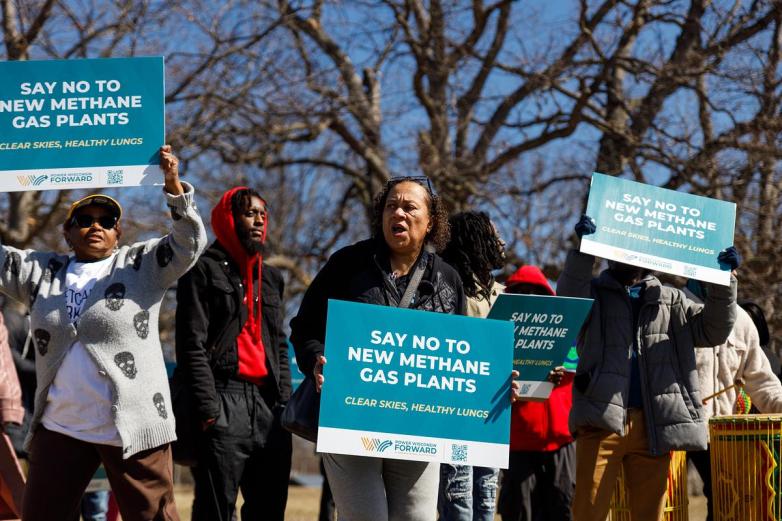 People hold signs reading “SAY NO TO NEW METHANE GAS PLANTS” outdoors with leafless trees in the background.
