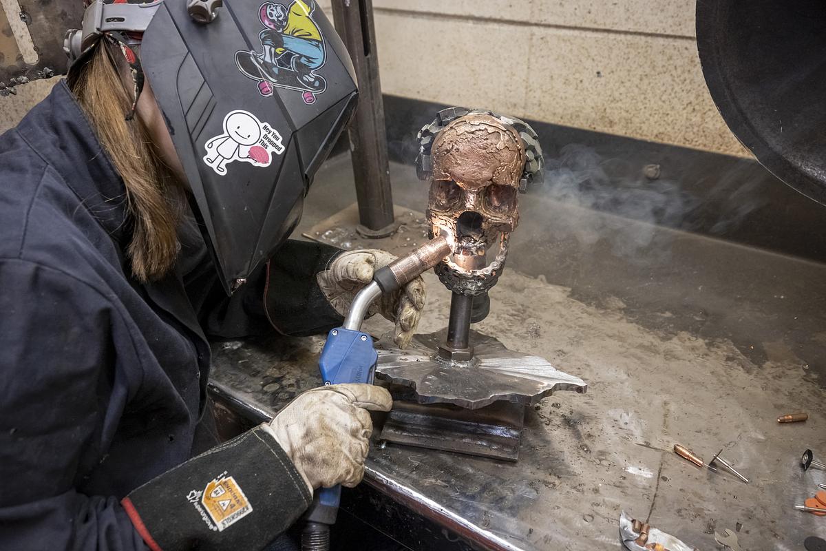 A person wearing gloves and a welding helmet holds a torch next to a metal skull mounted on a stand on a workbench, with smoke rising.