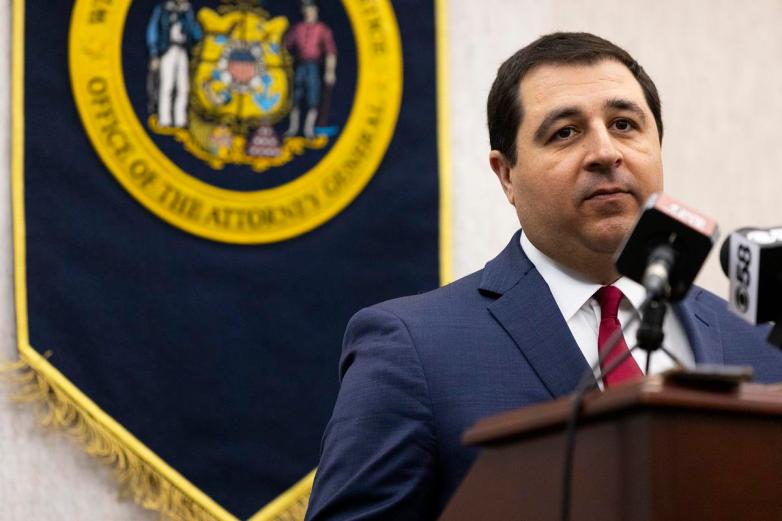 A person stands at a podium near microphones with a banner behind them displaying the Wisconsin state seal and the words 