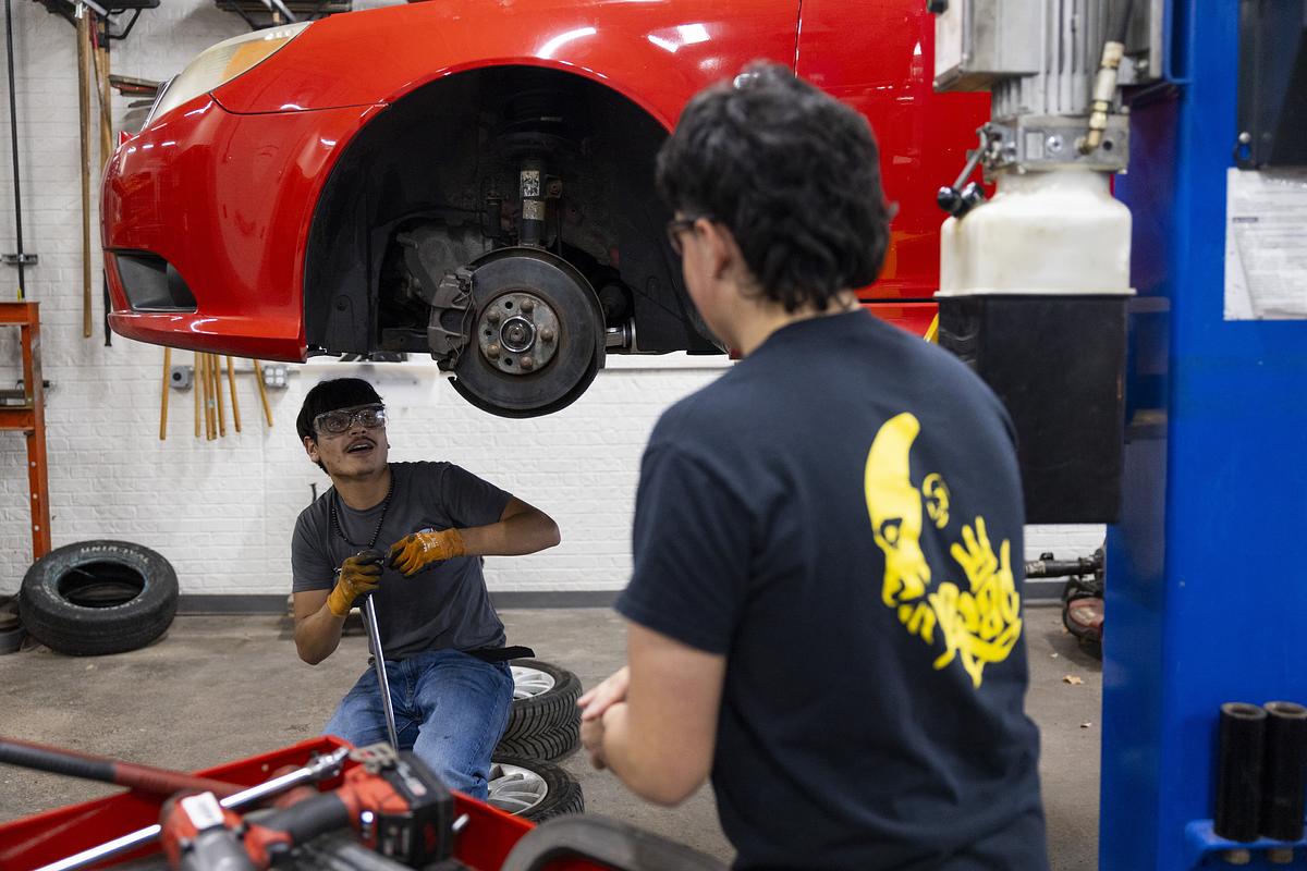 A person crouches under a raised red car and holds a tool while another person stands nearby, with loose tires and equipment on the floor around them.