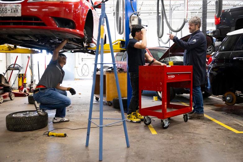 One person kneels under a raised red car, and two other people stand by a red tool cart, with one pointing a finger, in a big room with equipment and hoses visible.