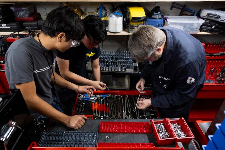 Three people wearing safety glasses look at wrenches and other hand tools in an open red tool drawer, with shelves of equipment and containers behind them.