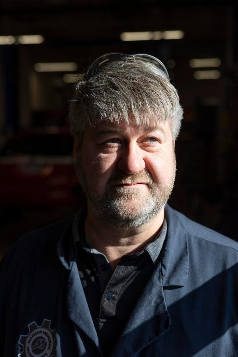 A person stands in bright light with safety glasses resting on the person's head, wearing a dark collared shirt and a jacket with a circular gear-shaped logo on the chest.