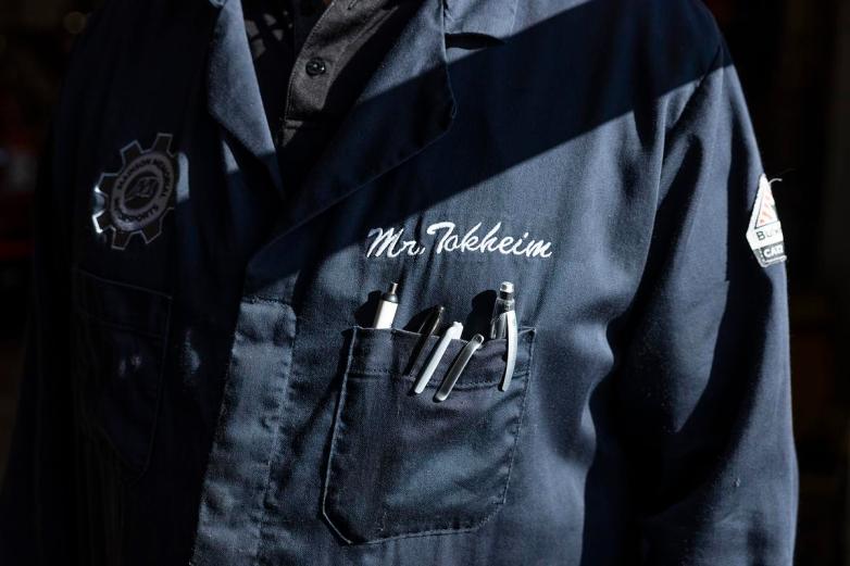 A dark jacket with a gear-shaped logo on one side and “Mr. Tokheim” stitched on the other, with pens and tools visible in the chest pocket.