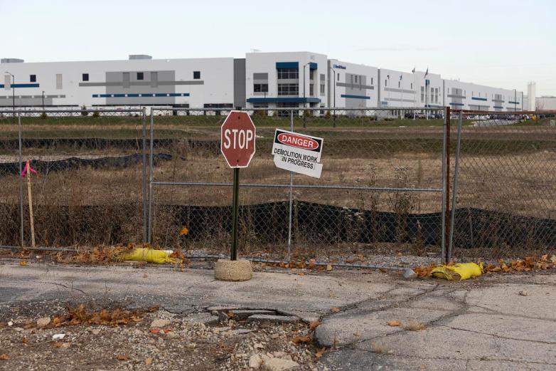 A chain-link fence, a “STOP” sign and a tilted “DANGER Demolition Work in Progress” sign stand in front of an open lot with a large industrial building in the background.