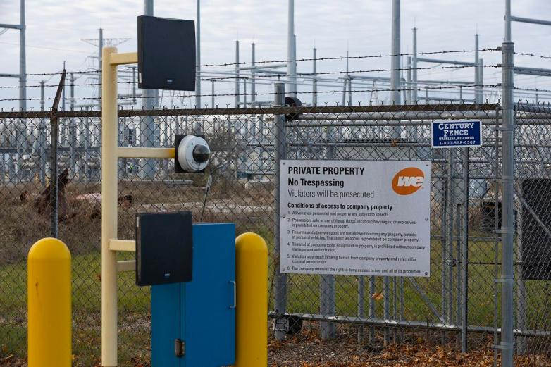 A barbed-wire fence with security cameras and signs reading “PRIVATE PROPERTY No Trespassing Violators will be prosecuted” stands in front of electrical equipment.