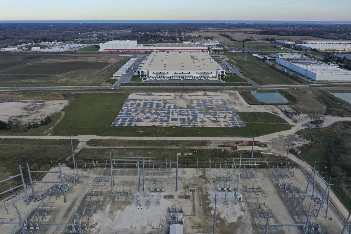 An aerial view of an electrical facility in the foreground. Beyond it are large industrial buildings, open fields and a rectangular patch of ground covered with blue sections.