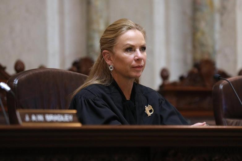 A person in a dark robe sits at a wooden bench with a microphone nearby and out-of-focus details in the background.