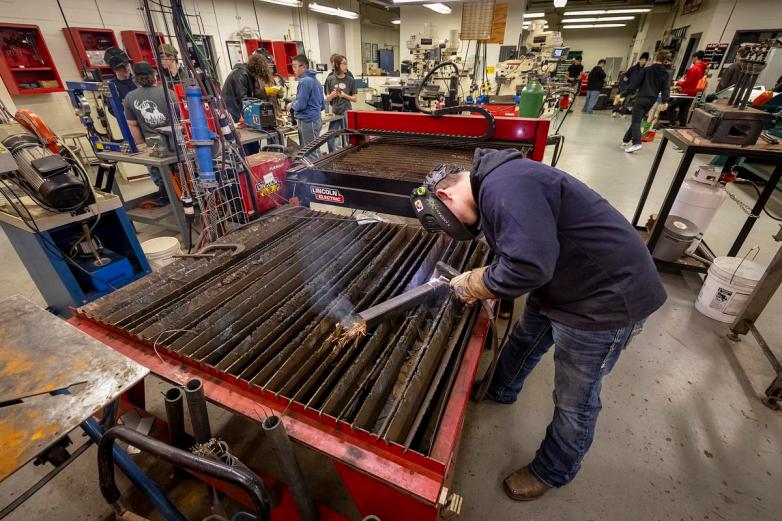 A person wearing gloves and a welding helmet uses a torch on a metal sheet atop a large table, with sparks flying and several other people and machines visible in a workshop.