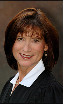 A person with short brown hair wearing a dark garment with a white collar looks toward the camera.