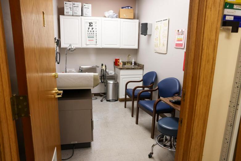 An exam room contains a padded exam table, two blue chairs, a sink with supplies, wall cabinets, medical posters, and equipment visible through an open door.