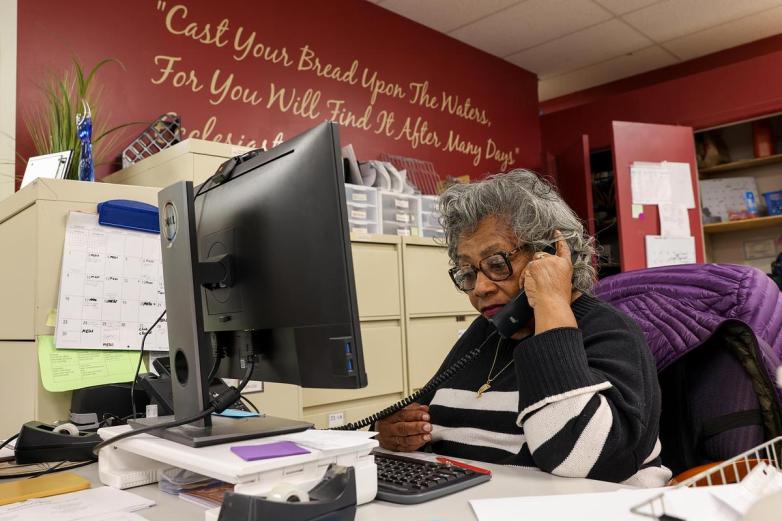 A person sits at a desk while holding a phone beside a computer monitor, with papers, office supplies, filing cabinets, and wall text in the background.
