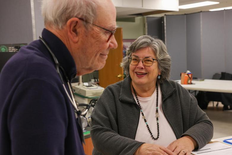 A person smiles and sits at a table across from another person wearing a stethoscope, with office equipment and partitions in the background.
