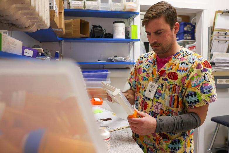 A person wearing a colorful patterned top holds a pill-counting tray while standing at a counter with medication bottles and shelves of supplies.