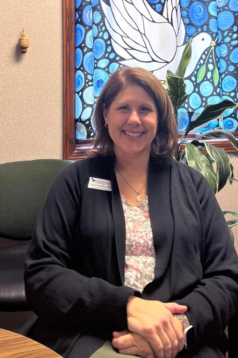 A person sits in a chair wearing a name badge, with patterned blue and white artwork featuring a dove on the wall behind.