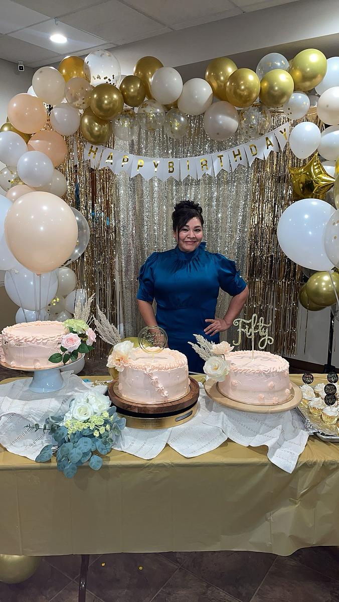 A person stands behind a table with three pink decorated cakes, surrounded by balloons, floral arrangements and a banner reading 
