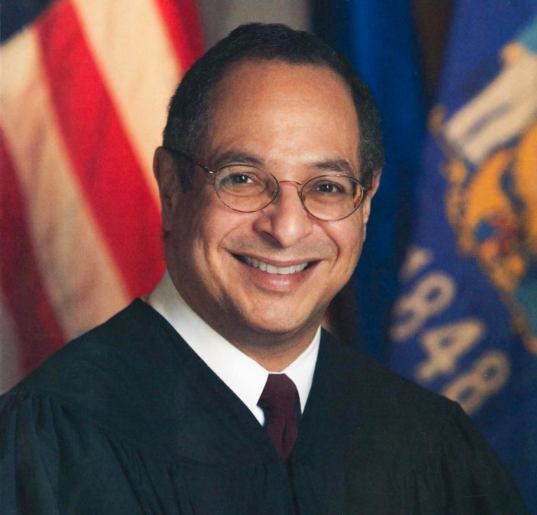 A person wearing glasses and a dark garment with a white collar and tie faces the camera with blurred flags in the background.