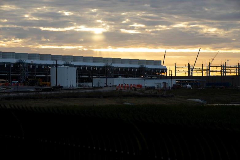 A large industrial building with rows of rooftop units stands behind construction barriers and cranes as sunlight breaks through clouds near the horizon.