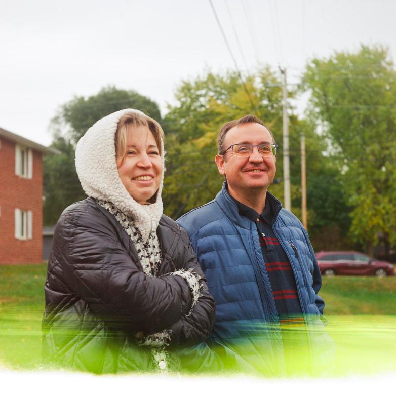 Two people stand outdoors wearing jackets, smiling and looking ahead, with trees, a grassy area and a brick building visible in the background.