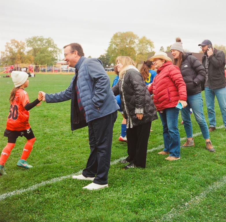 People stand in a line on a grassy field, exchanging high fives with soccer players in orange and blue uniforms.