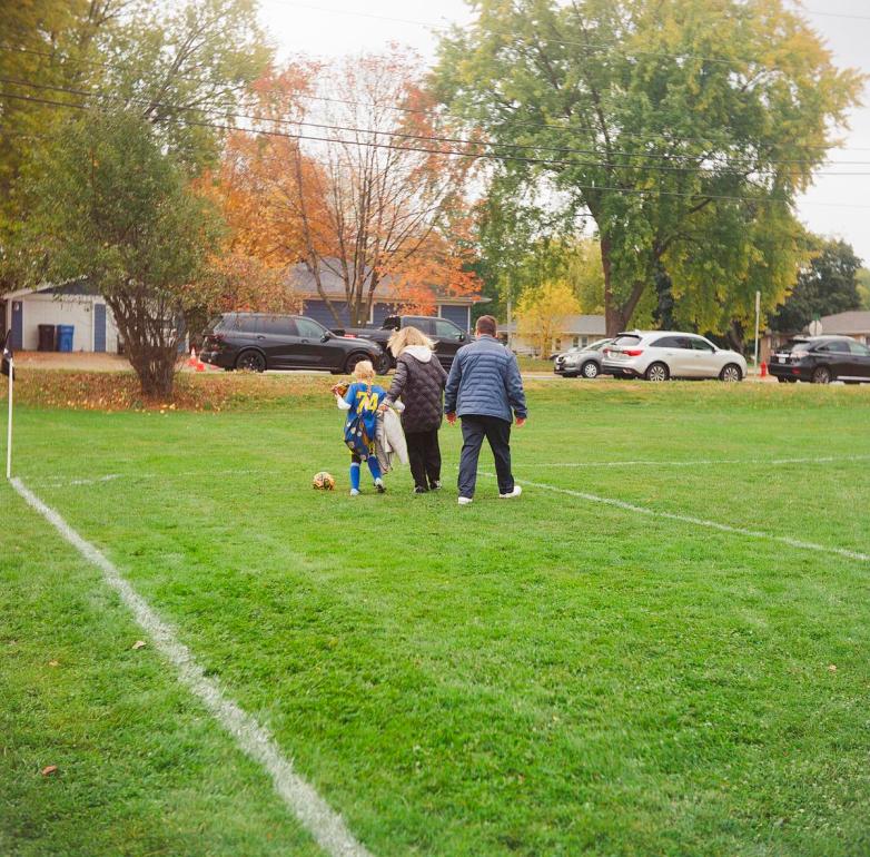 Three people are seen from behind walking across a grassy soccer field, with one wearing a blue jersey numbered “74” and carrying a bag, as a soccer ball rests nearby with parked cars in the background.