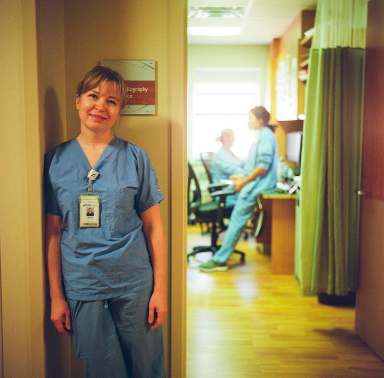 A person wearing blue scrubs and an ID badge stands beside a doorway, with two other people in scrubs seated at desks in a room behind the open door.